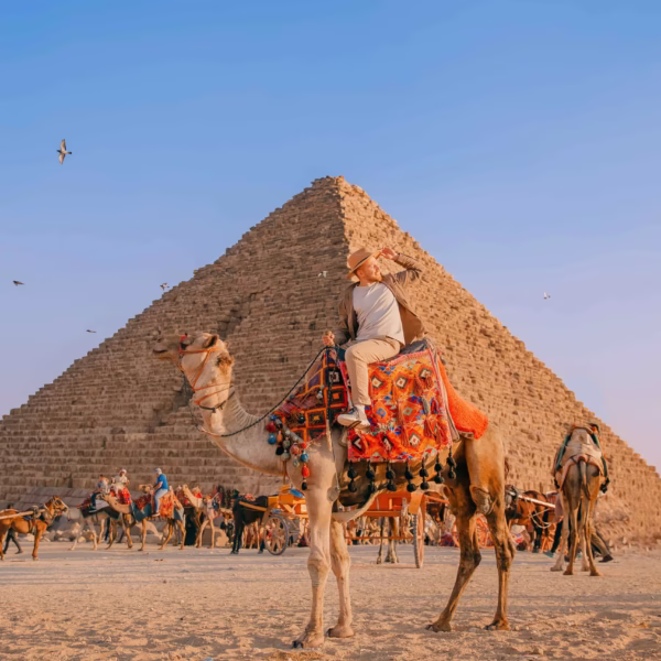 A traveler wearing a hat and light-colored clothing riding a camel with a traditional colorful saddle, positioned in front of a majestic ancient Egyptian pyramid under a clear blue sky.