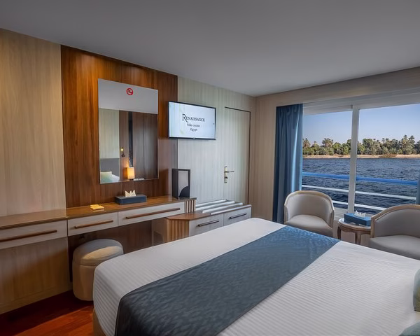 Modern Nile cruise suite with a double bed, blue runner, and floor-to-ceiling river views for a luxury Egypt tour.