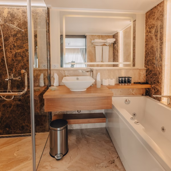 A luxury ship bathroom featuring a walk-in shower with brown marble walls, a white vessel sink on a wooden counter, and a white whirlpool bathtub, complemented by a large backlit mirror.