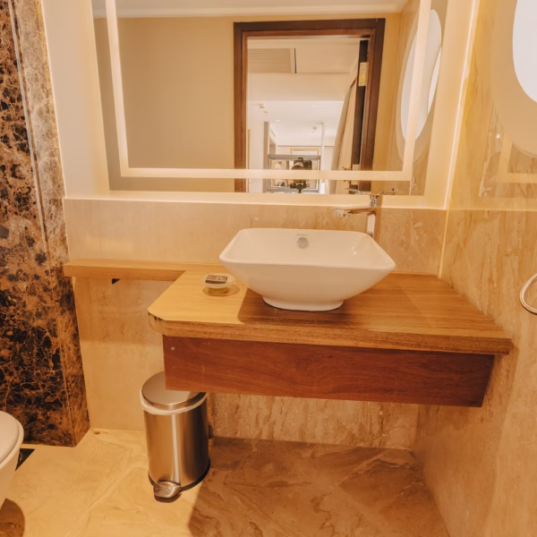 A modern bathroom vanity featuring a square white vessel sink on a wooden counter, a large backlit rectangular mirror, and a stainless steel step-trash can against marble-tiled walls