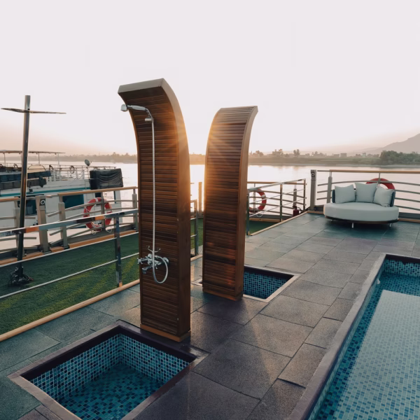 Two modern wooden outdoor showers on a cruise ship sun deck, positioned next to a blue tiled swimming pool with a scenic view of the Nile at sunset.