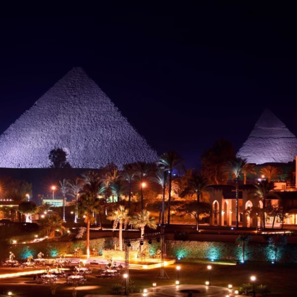 Night view of the illuminated Great Pyramids of Giza with a luxury resort and palm trees in the foreground.