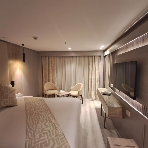 A modern luxury cruise cabin featuring a large bed with patterned pillows, a wall-mounted flat-screen TV on a backlit wood-slat panel, and contemporary artwork, with a doorway leading to a walk-in closet area.