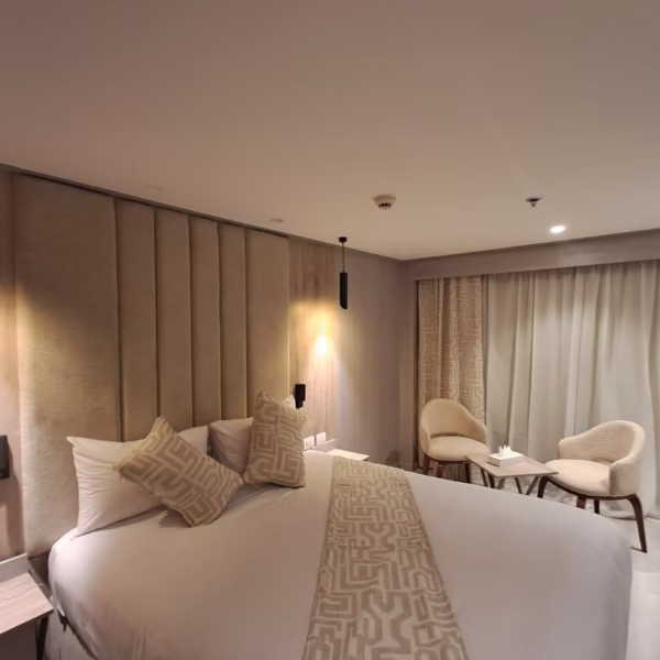 A modern luxury cruise cabin featuring a large bed with patterned pillows, a wall-mounted flat-screen TV on a backlit wood-slat panel, and contemporary artwork, with a doorway leading to a walk-in closet area.