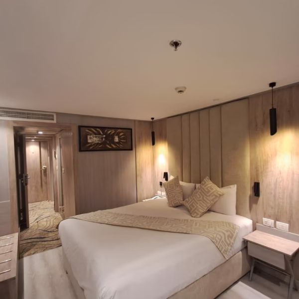 A modern luxury cruise cabin featuring a large bed with patterned pillows, a wall-mounted flat-screen TV on a backlit wood-slat panel, and contemporary artwork, with a doorway leading to a walk-in closet area.