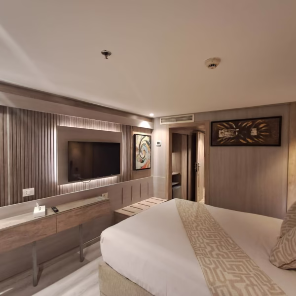 A modern luxury cruise cabin featuring a large bed with patterned pillows, a wall-mounted flat-screen TV on a backlit wood-slat panel, and contemporary artwork, with a doorway leading to a walk-in closet area.