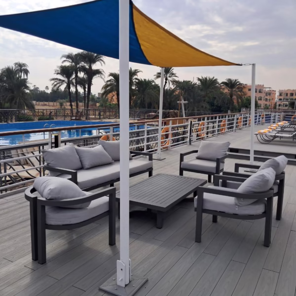 A luxury Nile cruise sun deck lounge area featuring modern grey cushioned outdoor sofas and chairs around a low coffee table, sheltered by a blue and yellow triangular sun shade, with a swimming pool and tropical shoreline in the background.