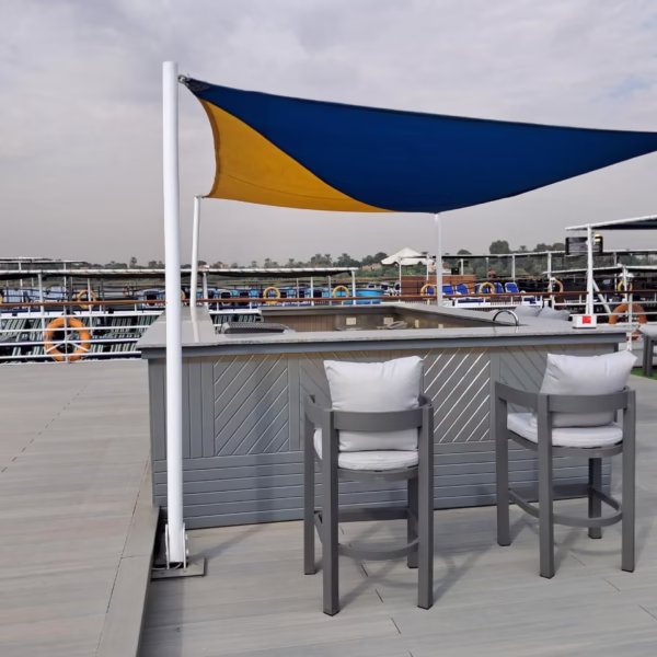 An outdoor sun deck bar on a luxury Nile cruise ship featuring a modern grey bar counter, two elegant high-back bar stools with grey cushions, and a stylish blue and yellow sun shade overlooking the river.