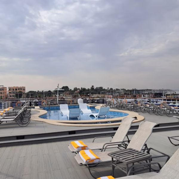 The sun deck of a luxury Nile cruise ship featuring a refreshing plunge pool with in-water lounge chairs, surrounded by comfortable sunbeds with yellow-striped towels and panoramic views of the riverbanks.