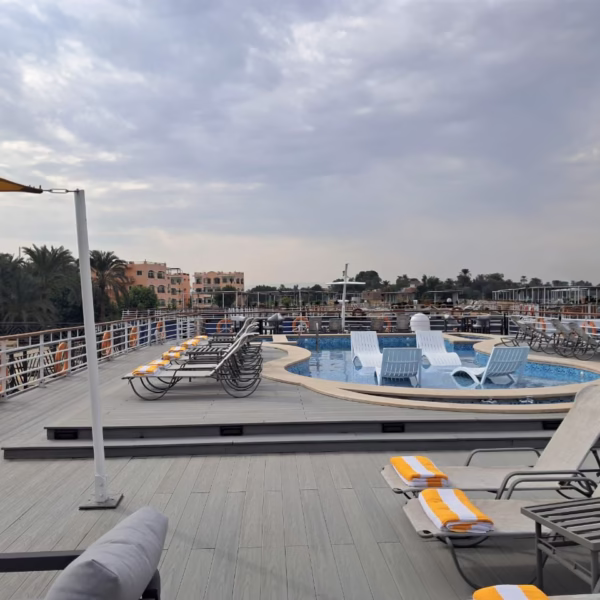 The sun deck of a luxury Nile cruise ship featuring a refreshing plunge pool with in-water lounge chairs, surrounded by comfortable sunbeds with yellow-striped towels and panoramic views of the riverbanks.
