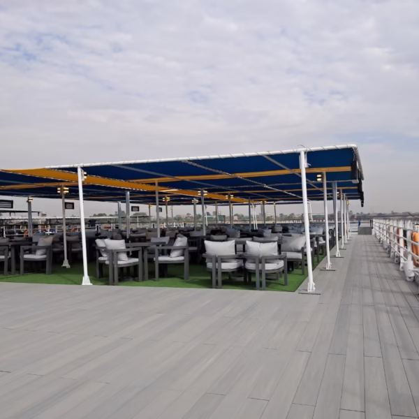 A spacious sun deck on a Nile cruise ship featuring a large shaded lounge area with grey cushioned seating under a blue and yellow canopy, set upon green artificial turf and grey wood-textured decking.