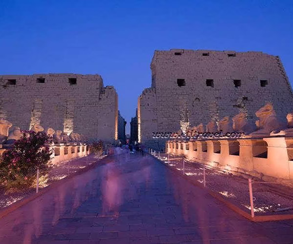 A wide night view of the illuminated Avenue of Sphinxes in Luxor, Egypt, featuring long rows of ancient stone statues leading toward the temple with bright spotlights and a church visible in the background.