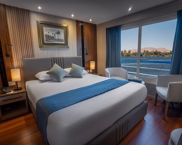 Luxury Nile cruise cabin with a queen bed, blue runner, and a large window facing the river for an Egypt tour.
