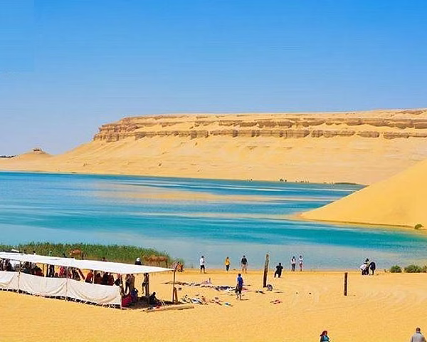 Tourists relax on a sandy beach by a vibrant blue lake in the Fayoum desert, a unique Egypt travel destination.